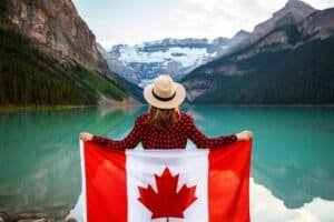Woman Wearing Red and Black Checkered Shirt and Beige Fedora Hat Holding Canada Flag Looking at Lake Louise Canada Is Canada Expensive to Visit? Canada Travel Cost & Budget Guide 2026 Woman Holding Canadian Flag Looking at Lake Louise, Canada